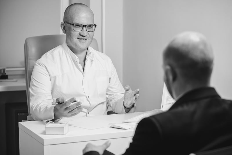 A man in a white lab coat sitting at a desk, gesturing with his hands while speaking to another man seen from the back.