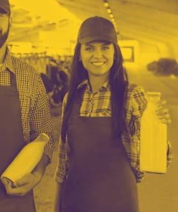 A smiling woman in a plaid shirt, apron, and cap holds a bottle of milk