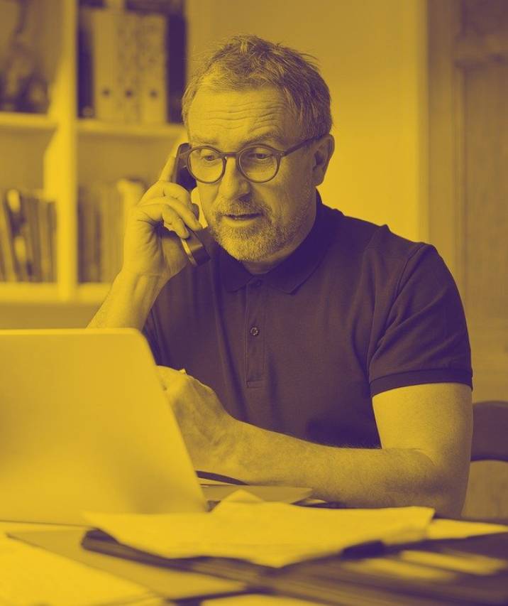 A middle-aged man with glasses and a beard talking on a telephone while looking at a laptop