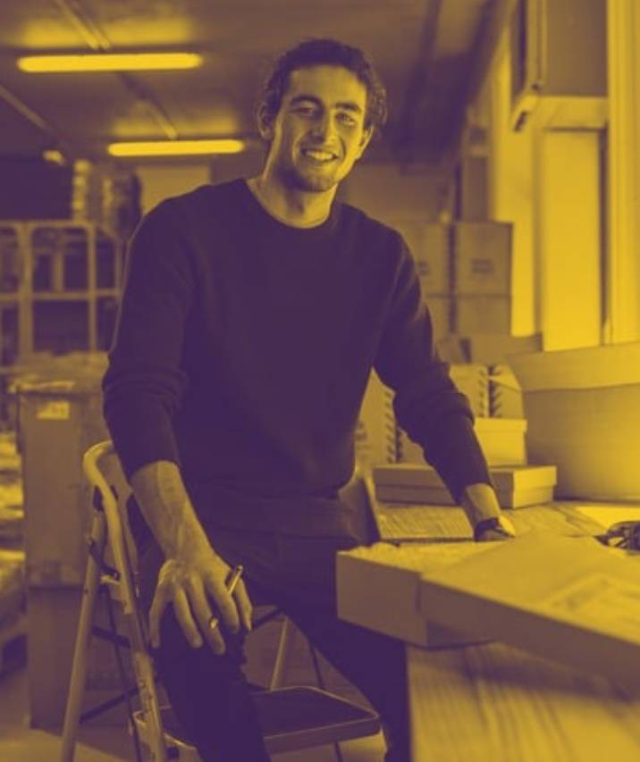A smiling man in a dark sweater sitting at a desk with boxes and packing materials in a yellow-tinted warehouse or shipping room.
