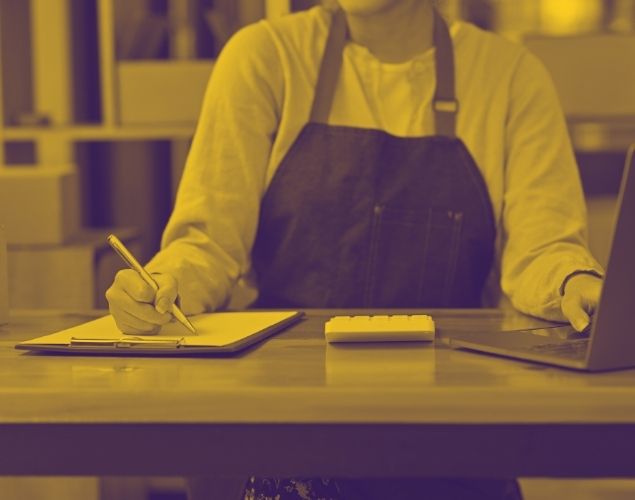 A person wearing an apron sitting at a desk and writing on a clipboard while using a laptop